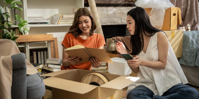 Two women sitting on a checkered rug in a room with boxes and books. One of the women is holding a cell phone
